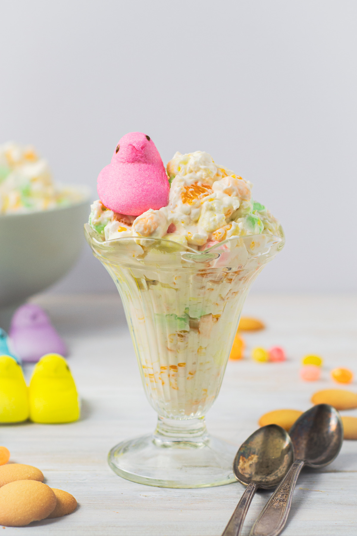 A glass dessert dish filled with creamy fruit fluff, layered with colorful ingredients, and topped with a pink Peeps chick, with vintage spoons.